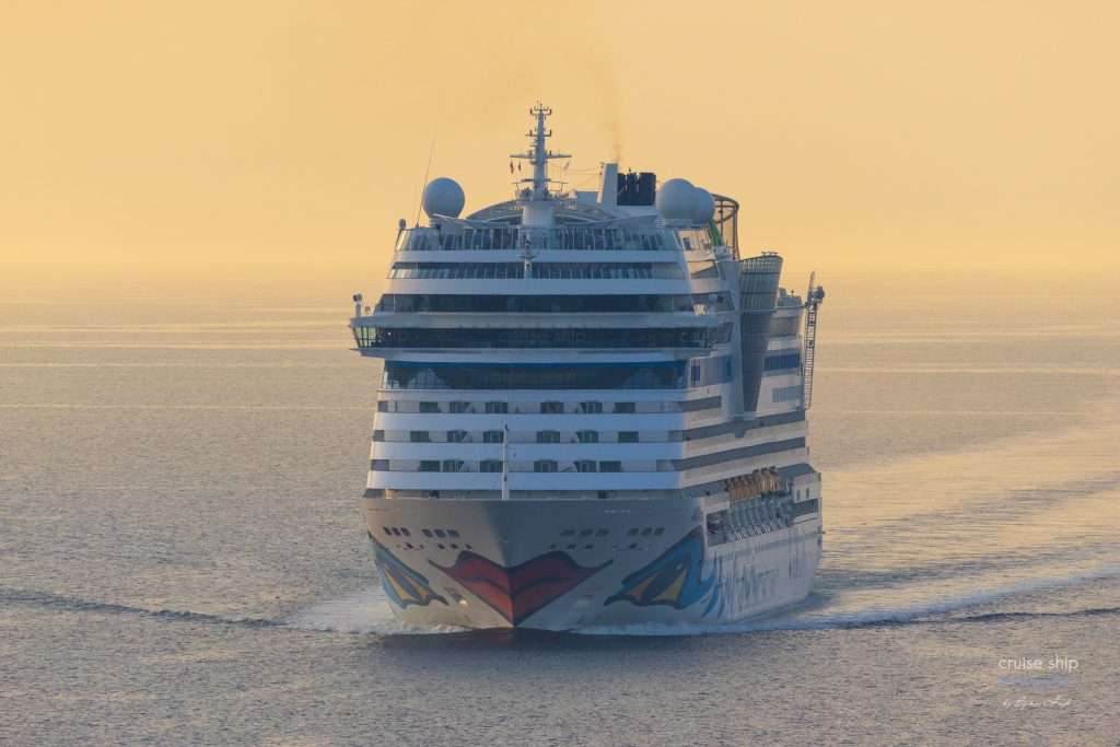 Kreuzfahrtschiff AIDAmar im goldenen Abendlicht auf dem Meer. Das Meer ist glatt und die Luftaufnahme zeigt das Schiff schräg von vorne